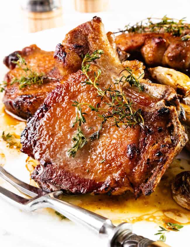 A close-up of a savory, pan-seared brined pork chop garnished with fresh herbs on a white plate. The chop is crispy and golden-brown with a juicy texture. In the background, two salt and pepper shakers are slightly blurred. The dish is served with a fork and knife.