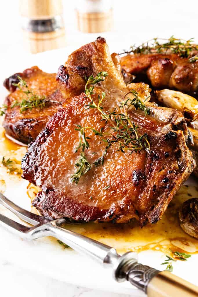 A close-up of a savory, pan-seared brined pork chop garnished with fresh herbs on a white plate. The chop is crispy and golden-brown with a juicy texture. In the background, two salt and pepper shakers are slightly blurred. The dish is served with a fork and knife.