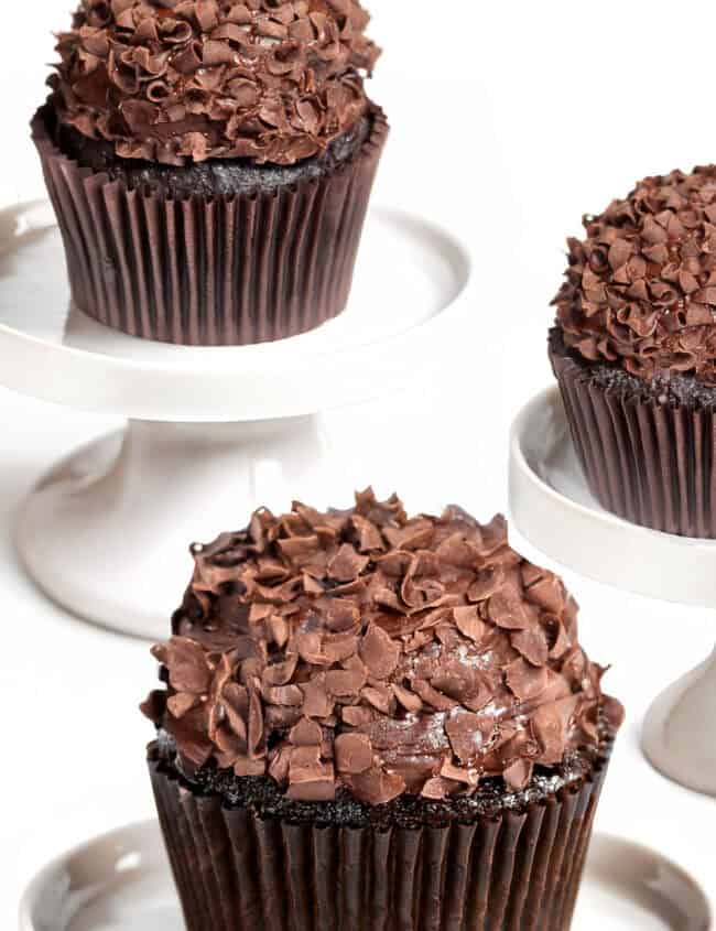 Three chocolate cupcakes, rich and indulgent, adorned with delicate chocolate shavings, are beautifully displayed on white pedestal stands against a pristine white background.