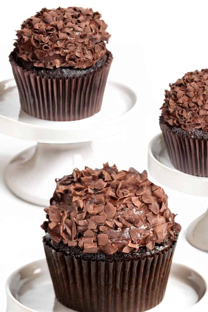 Three chocolate cupcakes, rich and indulgent, adorned with delicate chocolate shavings, are beautifully displayed on white pedestal stands against a pristine white background.