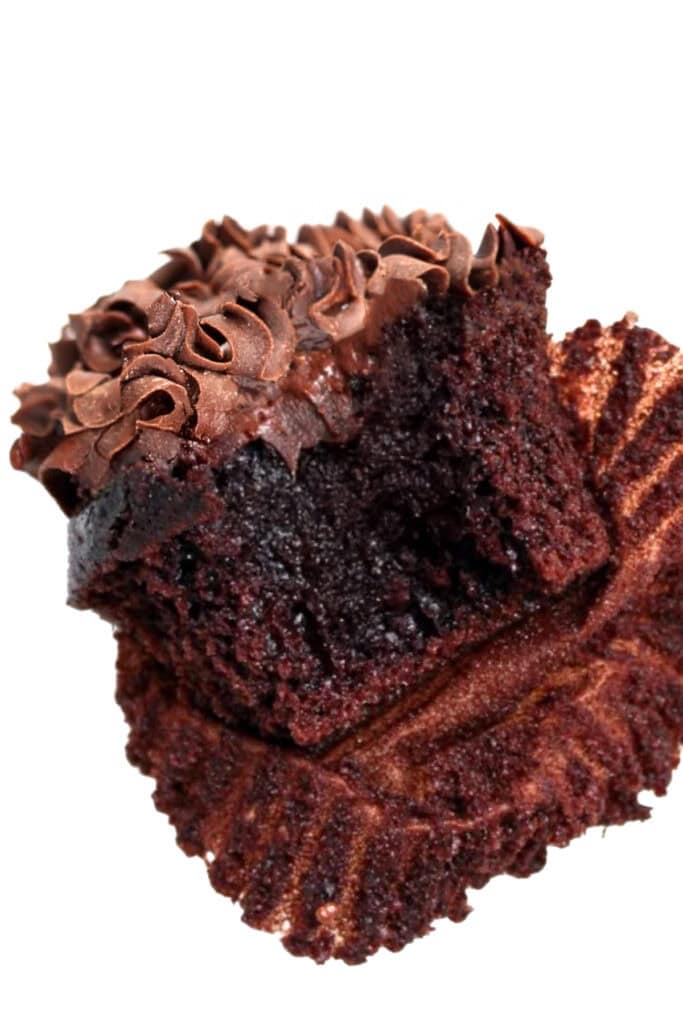 A chocolate cupcake with rich frosting is partially unwrapped and has a bite taken out, revealing its moist, dark interior. The delicate swirled peaks on the frosting make these chocolate cupcakes an irresistible treat.