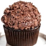 A luscious chocolate cupcake with creamy frosting and delicate chocolate shavings graces a white surface. Nestled in a dark brown paper liner, this delectable treat epitomizes the rich indulgence of classic Chocolate Cupcakes.