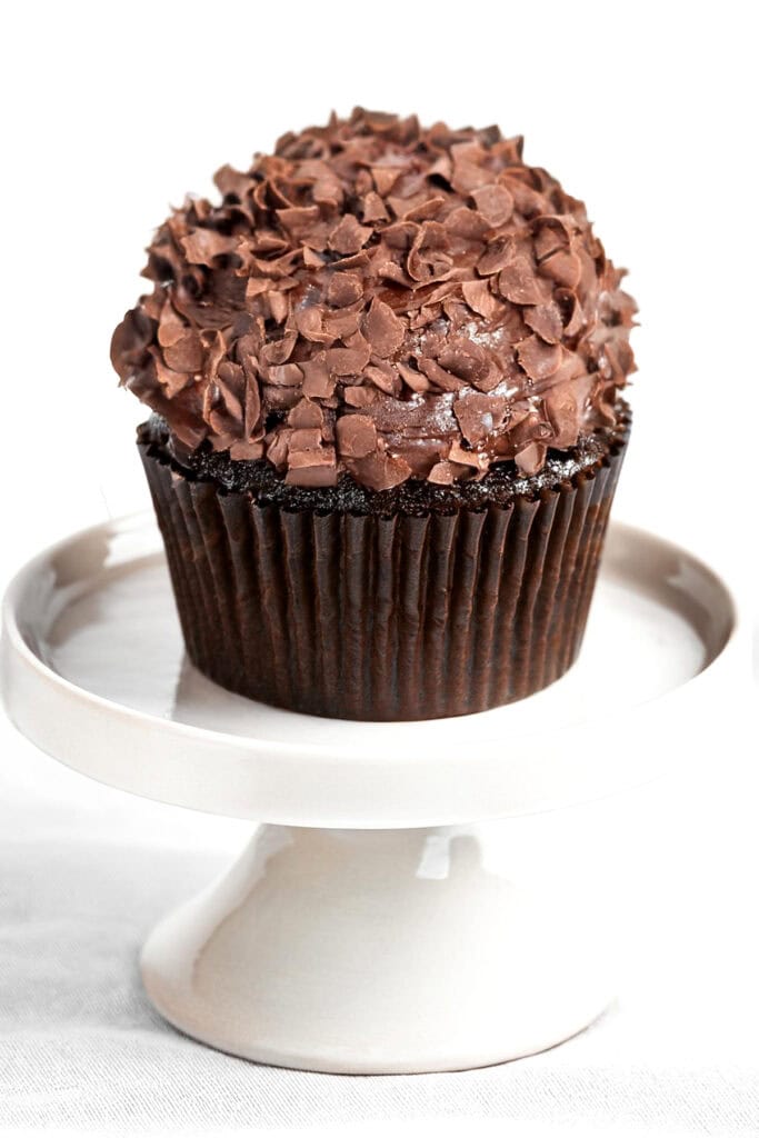 A luscious chocolate cupcake topped with a generous amount of chocolate shavings sits elegantly on a white ceramic pedestal stand. The textured surface and dark brown wrapper perfectly showcase the rich allure of chocolate cupcakes against the crisp white background.