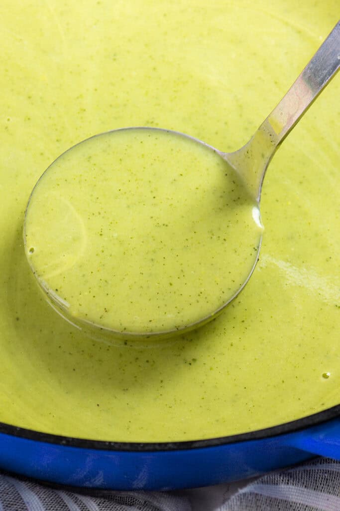 A Ladle of purified zucchini soup being held over a blue pot filled with soup.