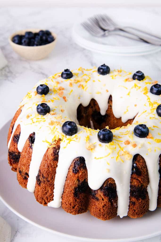 A close-up photo of a lemon blueberry cake on a plate. The cake is a light purple color with blueberries scattered throughout the inside. The top of the cake is drizzled with a white icing and sprinkled with cake crumbs.