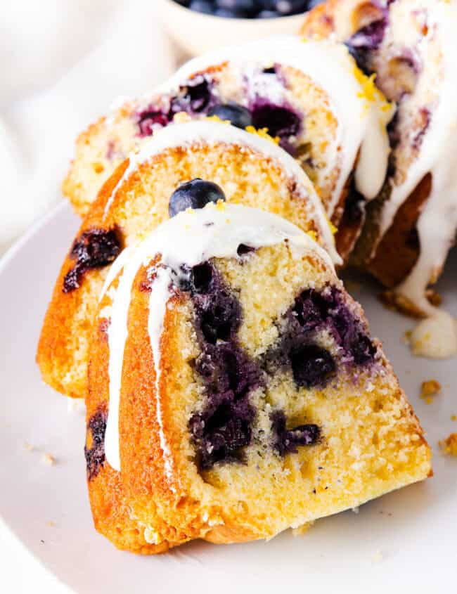 A sliced bundt cake drizzled with a white icing and sprinkled with lemon zest on a white plate. With blueberries visible throughout the center.