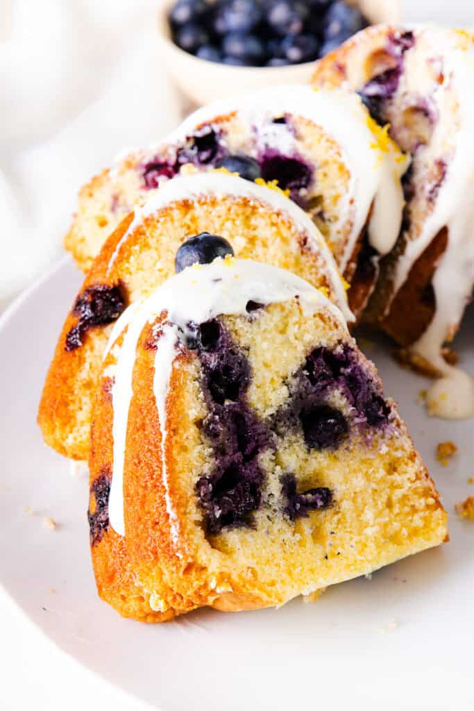 A sliced bundt cake drizzled with a white icing and sprinkled with lemon zest on a white plate. With blueberries visible throughout the center.