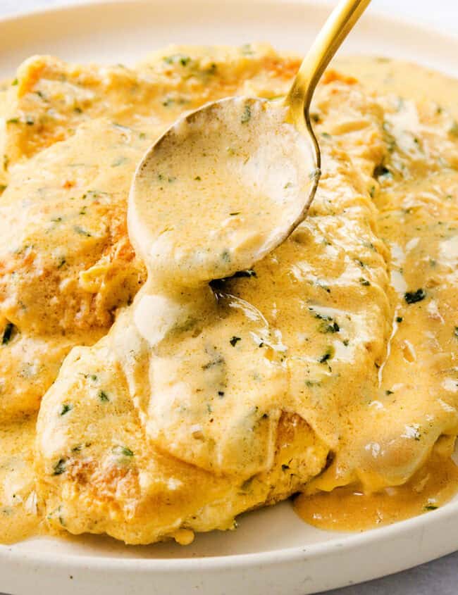 A plate of mustard chicken smothered in a creamy sauce, garnished with fresh herbs. A spoon drizzles more sauce over the dish, enhancing its rich flavor. The background is softly blurred, highlighting this delicious meal.