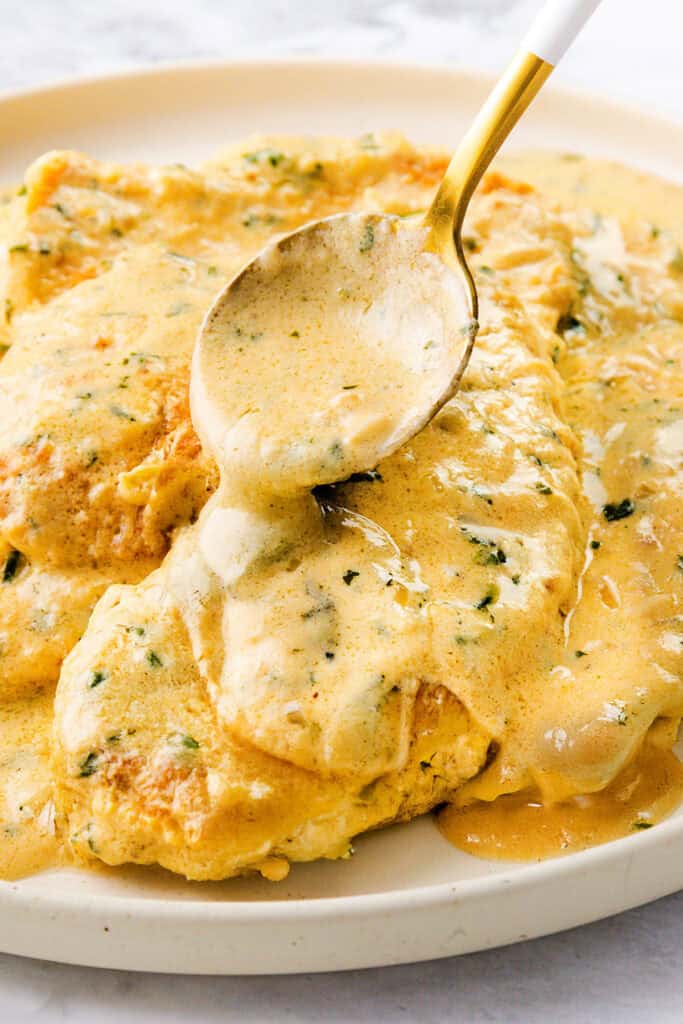 A plate of mustard chicken smothered in a creamy sauce, garnished with fresh herbs. A spoon drizzles more sauce over the dish, enhancing its rich flavor. The background is softly blurred, highlighting this delicious meal.