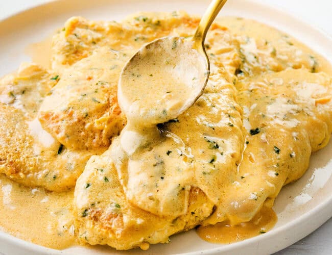 A plate of mustard chicken is being served with a spoon, featuring creamy, herb-infused goodness. The chicken is smothered in a rich, yellow mustard sauce with visible herbs, giving it an appetizing appearance.