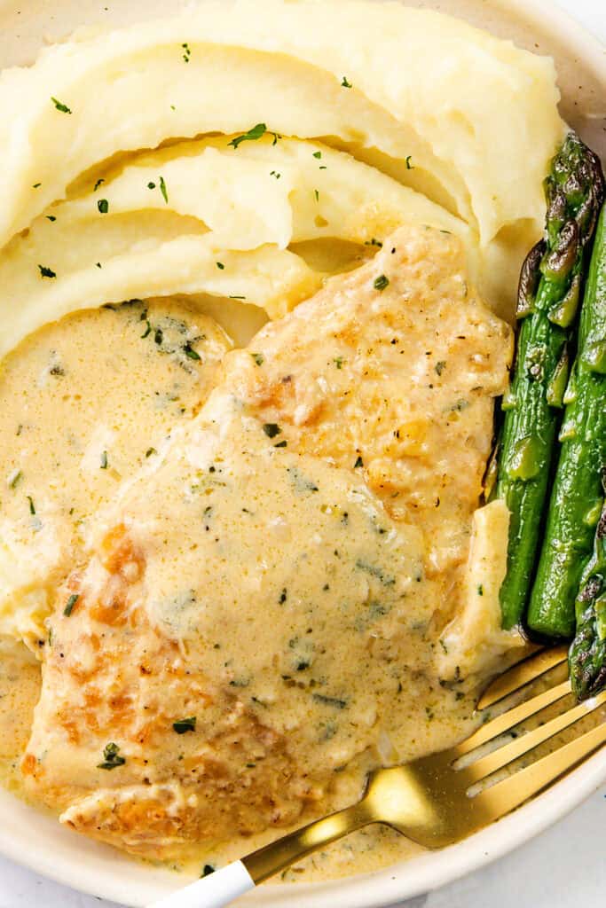 A plate of creamy mustard chicken in sauce, served alongside mashed potatoes and roasted asparagus. A fork rests on the dish.