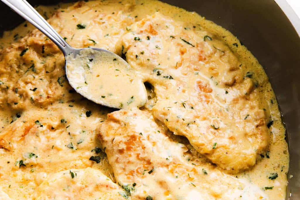 Mustard Chicken transforms a creamy chicken dish in a pan, with tender pieces of chicken breast smothered in a rich, herb-flecked mustard sauce. A silver spoon rests invitingly in the sauce, ready to serve.