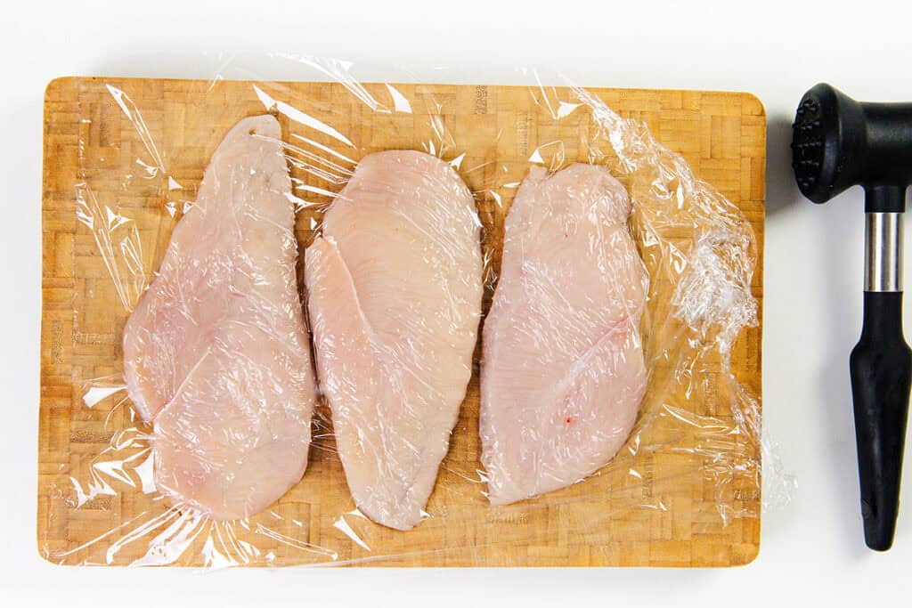 Three raw chicken breasts, soon to be transformed into delicious mustard chicken, are covered in clear plastic wrap on a wooden cutting board. A black meat tenderizer is ready at hand, poised for action next to the board on the right.