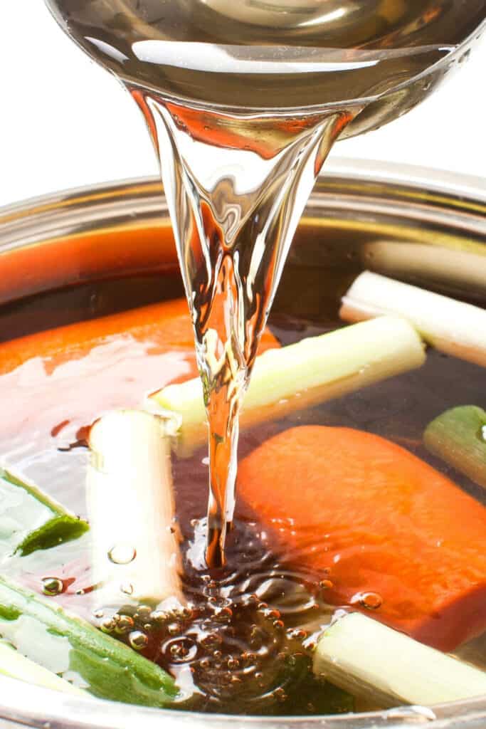 water being poured into a large pan with chopped vegetables and chicken carcass