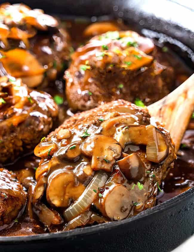 A skillet with juicy Salisbury steak patties topped with sautéed mushrooms, onions, and rich brown gravy. Fresh chopped herbs are sprinkled on top. A wooden spoon holds a single Salisbury steak patty, highlighting its tender, perfectly cooked texture.