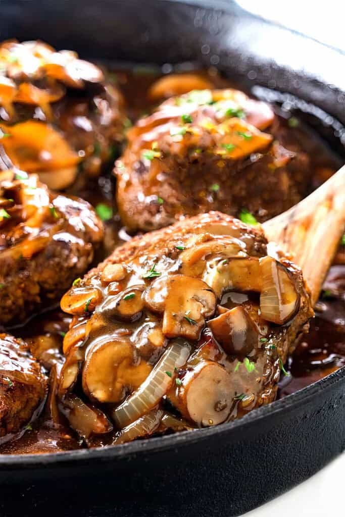 A skillet with juicy Salisbury steak patties topped with sautéed mushrooms, onions, and rich brown gravy. Fresh chopped herbs are sprinkled on top. A wooden spoon holds a single Salisbury steak patty, highlighting its tender, perfectly cooked texture.