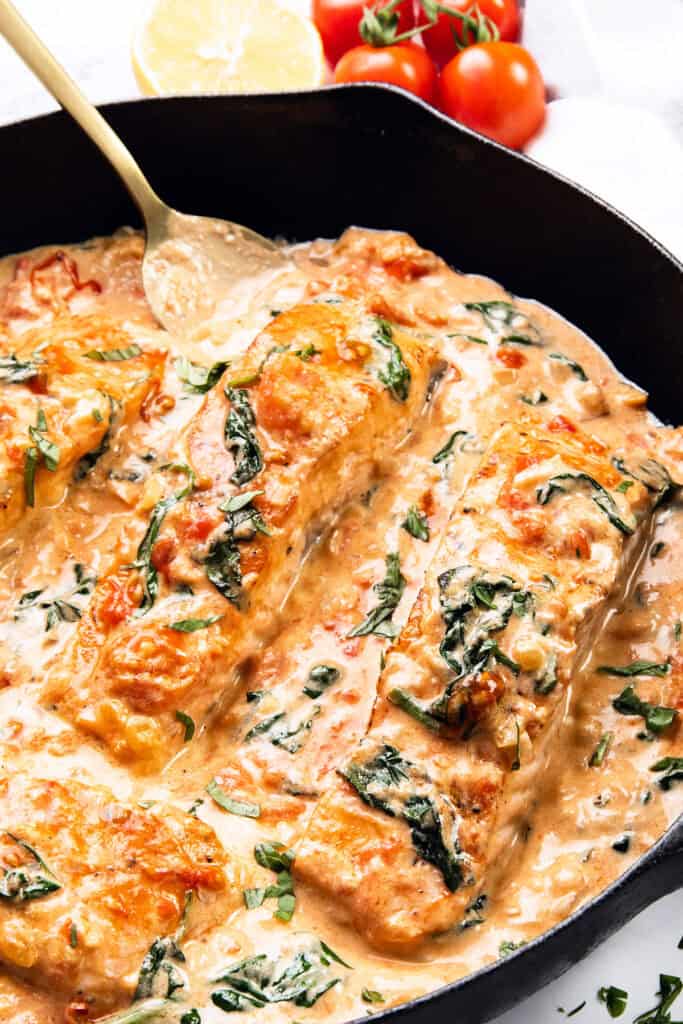 Tuscan Salmon fillets in a creamy sauce with spinach, tomatoes, and herbs simmer in a skillet. A spoon rests nearby, while fresh cherry tomatoes and a lemon half add vibrant touches in the background.