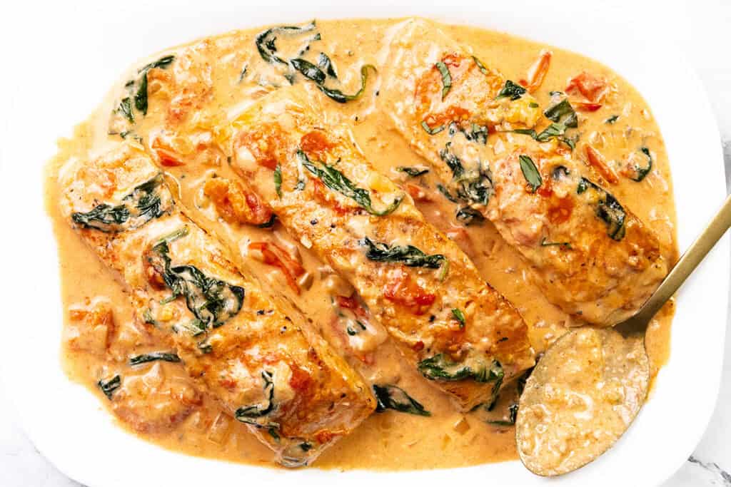 Three pieces of Tuscan Salmon in a creamy tomato and spinach sauce, garnished with herbs, served on a white plate. A spoon rests in the sauce beside the salmon.