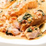 A dish of Tuscan Salmon fillets in a creamy sauce with spinach and tomatoes, served on a white plate. A fork is taking a piece of the salmon, revealing its flaky texture.