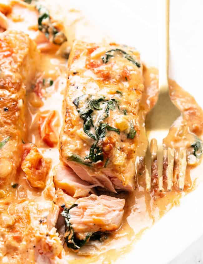 A close-up of Tuscan Salmon with creamy sauce, spinach, and tomatoes. The salmon is cooked to a tender texture, topped with a rich sauce and garnished with vegetables. A gold fork is on the plate beside the fish.