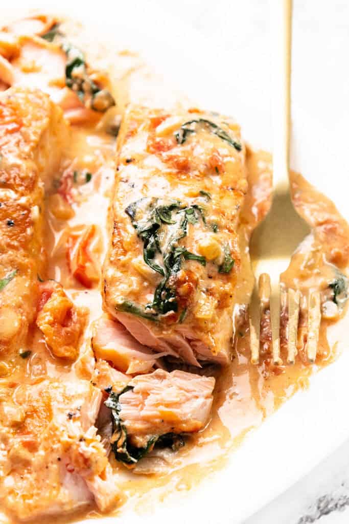 A close-up of Tuscan Salmon with creamy sauce, spinach, and tomatoes. The salmon is cooked to a tender texture, topped with a rich sauce and garnished with vegetables. A gold fork is on the plate beside the fish.