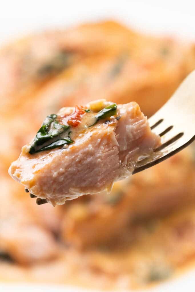 A fork holds a piece of Tuscan Salmon, cooked to perfection and topped with vibrant herbs and seasoning. The salmon appears tender and flaky, with green herbs and a touch of red seasoning adding to its allure. The softly blurred background enhances its savory appeal.