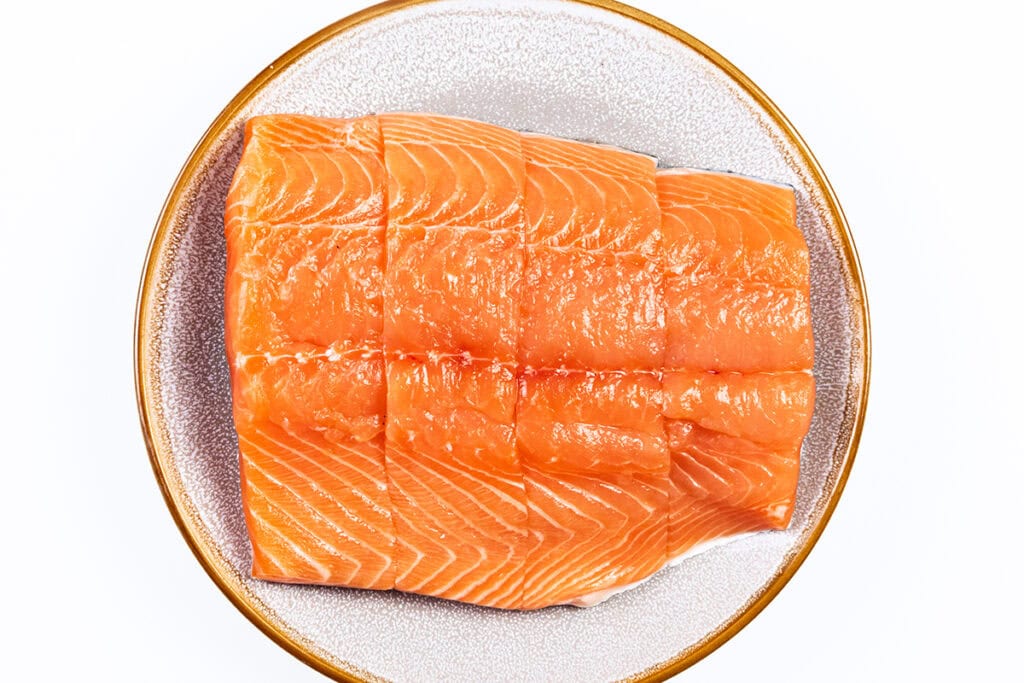 A plate with raw salmon fillet, divided into neat, rectangular pieces. The bright orange salmon, with visible marbling lines, is elegantly set on a circular plate with a golden rim.