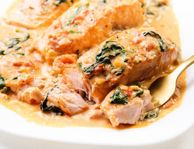 A dish of Tuscan Salmon fillets in a creamy sauce with spinach and tomatoes, served on a white plate. A fork is taking a piece of the salmon, revealing its flaky texture.