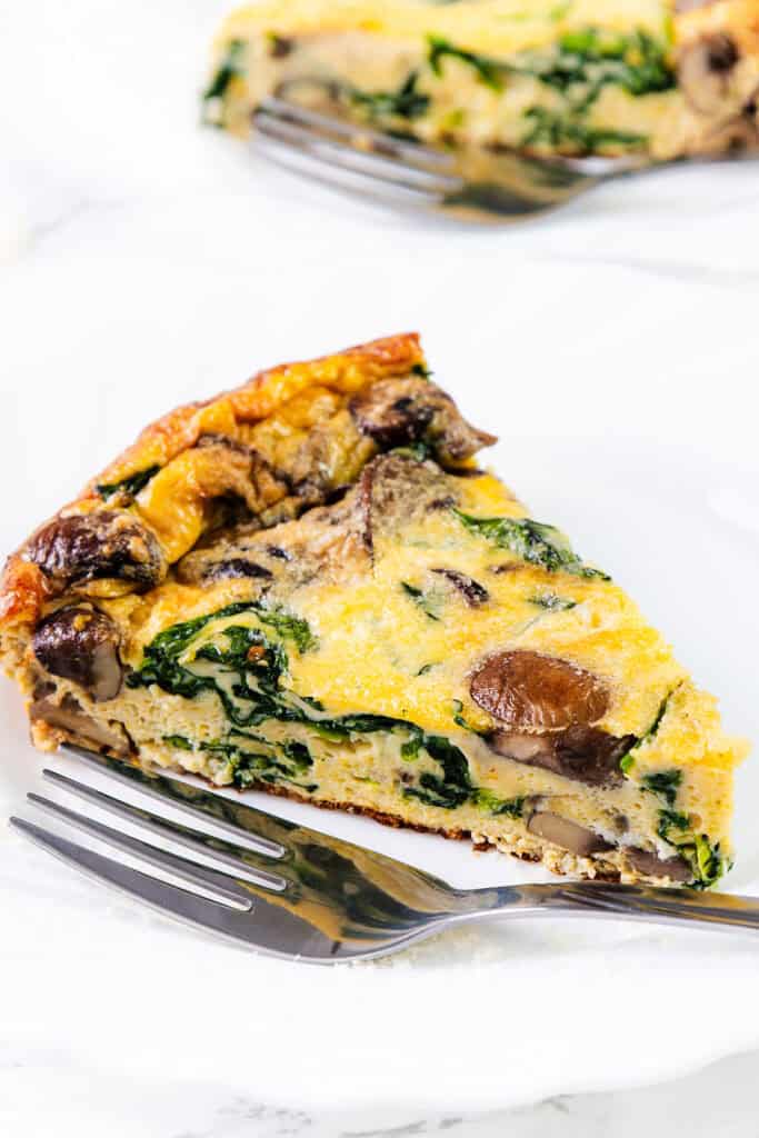 A close up image of a slice of Easy Spinach Mushroom Frittata on a white plate with a fork.