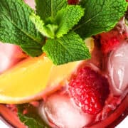 A refreshing Raspberry Mojito beverage garnished with a lemon wedge, fresh mint leaves, and a raspberry is shown over ice. The colorful drink is set against a white background, creating a vibrant and appealing presentation.