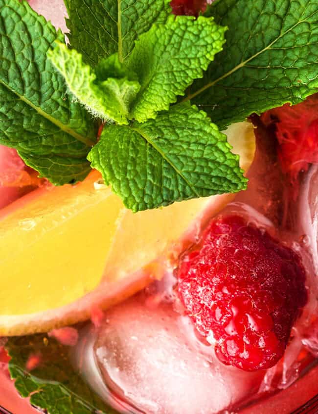 A refreshing Raspberry Mojito beverage garnished with a lemon wedge, fresh mint leaves, and a raspberry is shown over ice. The colorful drink is set against a white background, creating a vibrant and appealing presentation.