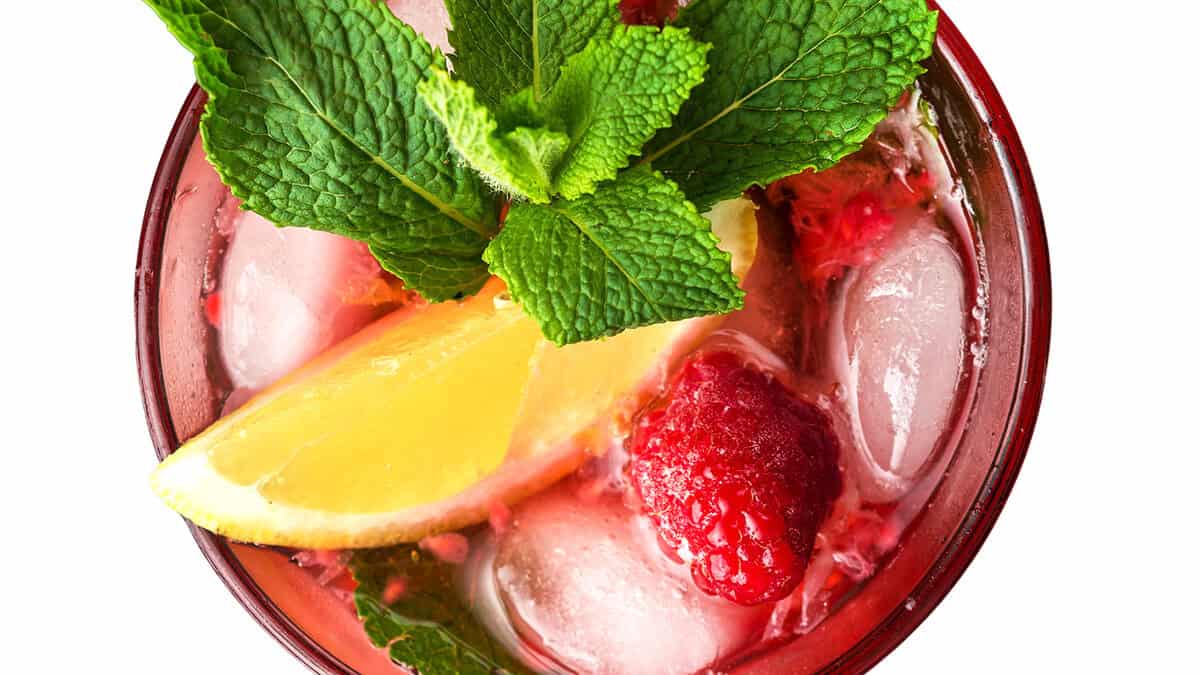 A refreshing Raspberry Mojito beverage garnished with a lemon wedge, fresh mint leaves, and a raspberry is shown over ice. The colorful drink is set against a white background, creating a vibrant and appealing presentation.