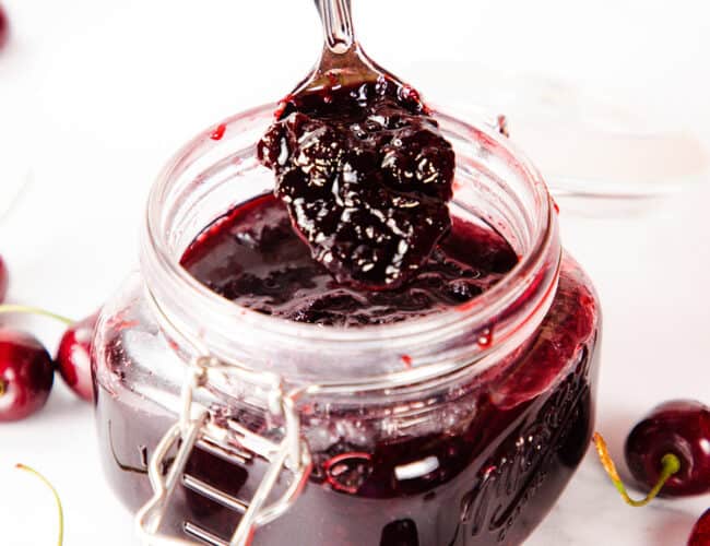 a spoon of cherry jam preserves over a full jar with fresh cherries all around it