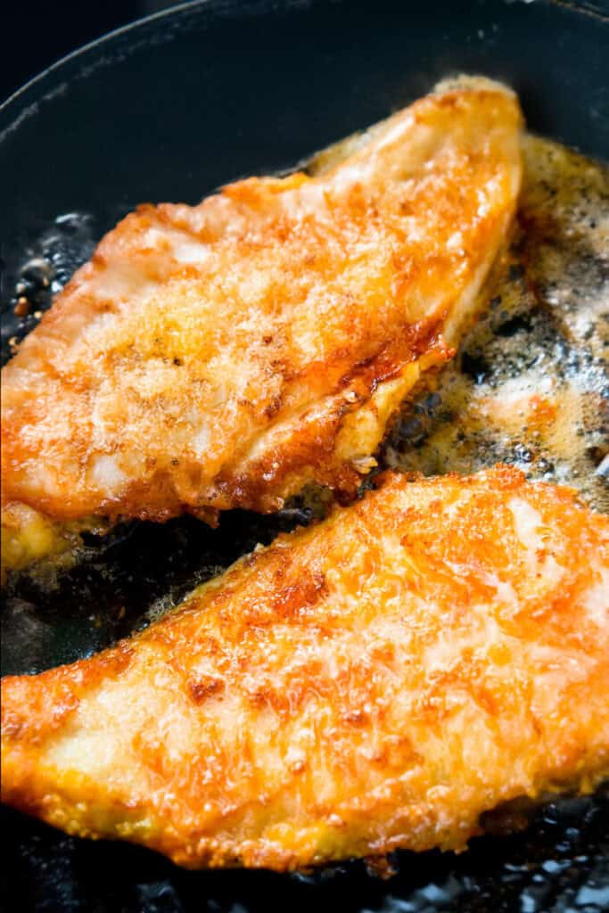 Two Super Crispy Pan Fried Fish fillets are sizzling in hot oil, their crispy, lightly seasoned crust.