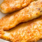 Golden-brown pan fried fish fillets are neatly arranged on a white plate, showcasing their crispy texture. A wedge of lemon is placed beside them, adding a splash of color to the dish.