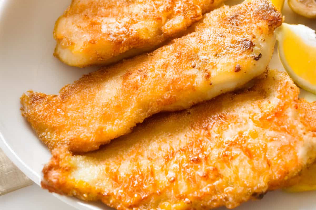 Golden-brown pan fried fish fillets are neatly arranged on a white plate, showcasing their crispy texture. A wedge of lemon is placed beside them, adding a splash of color to the dish.