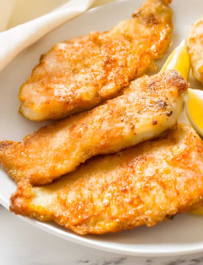 A close-up of a plate featuring three pieces of golden-brown pan fried fish, arranged in a slight overlap.