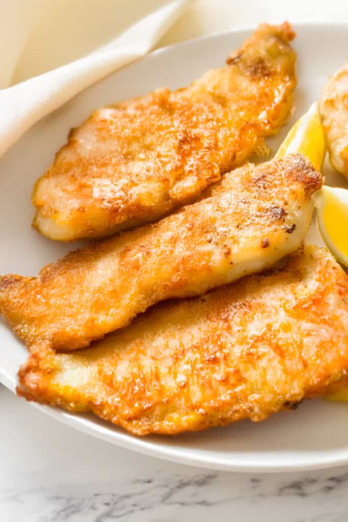 A close-up of a plate featuring three pieces of golden-brown pan fried fish, arranged in a slight overlap. 