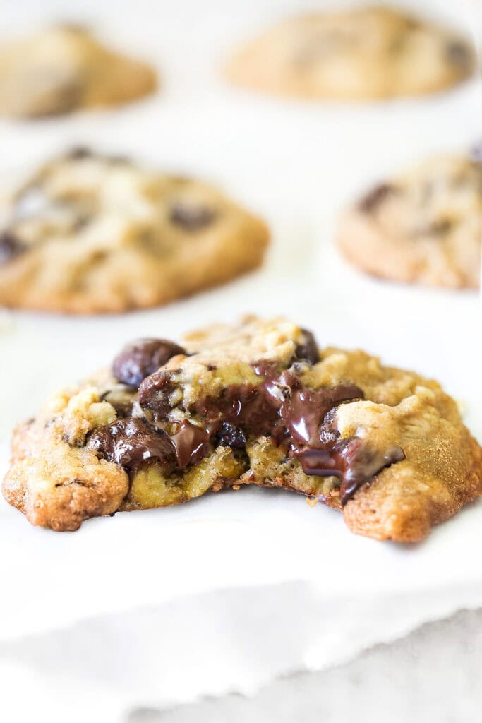 Close-up of a freshly baked, easy chocolate chip oatmeal cookie with a gooey, melted chocolate center, displayed on a white surface. More cookies are blurred in the background.