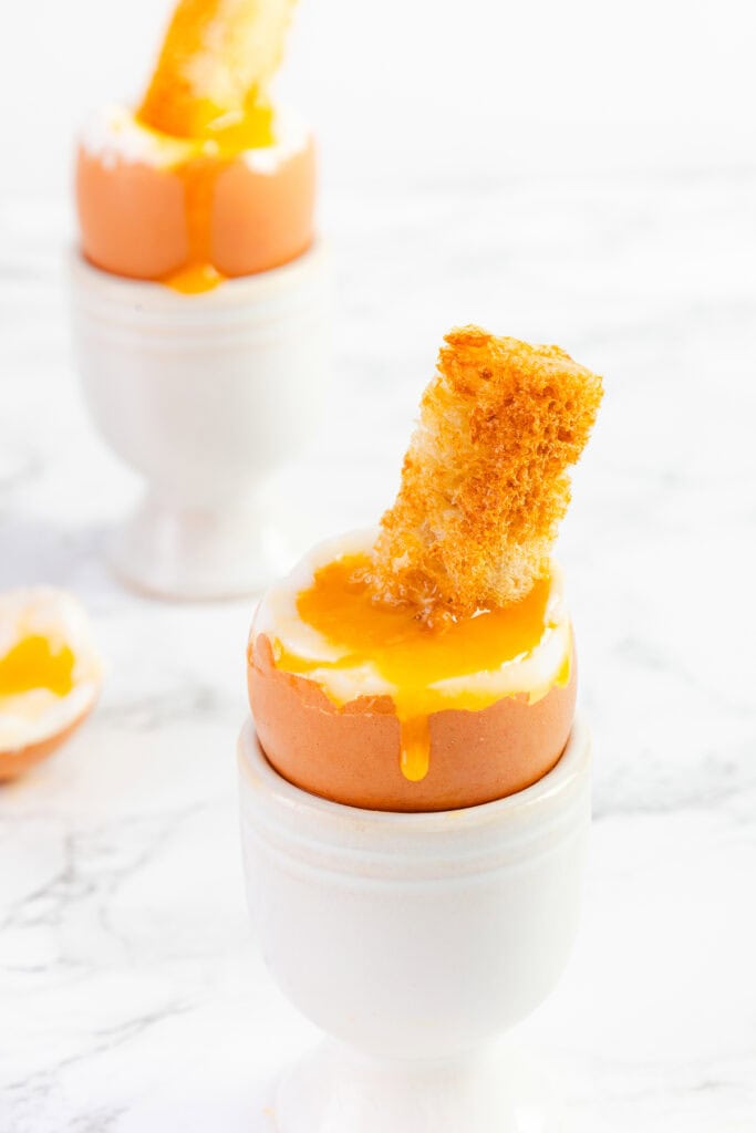 A soft-boiled egg sits in a white egg cup with a piece of toast dipped into the runny yolk, showing how to soft boil an egg perfectly. The toast is golden and crisp, and the yolk drips slightly down the side of the egg. Another similarly prepared egg is visible in the background.