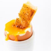 A soft-boiled egg with a runny yolk and a crispy, golden toast stick dipped into it, placed in a white egg cup against a plain white background captures the essence of how to soft boil an egg perfectly.