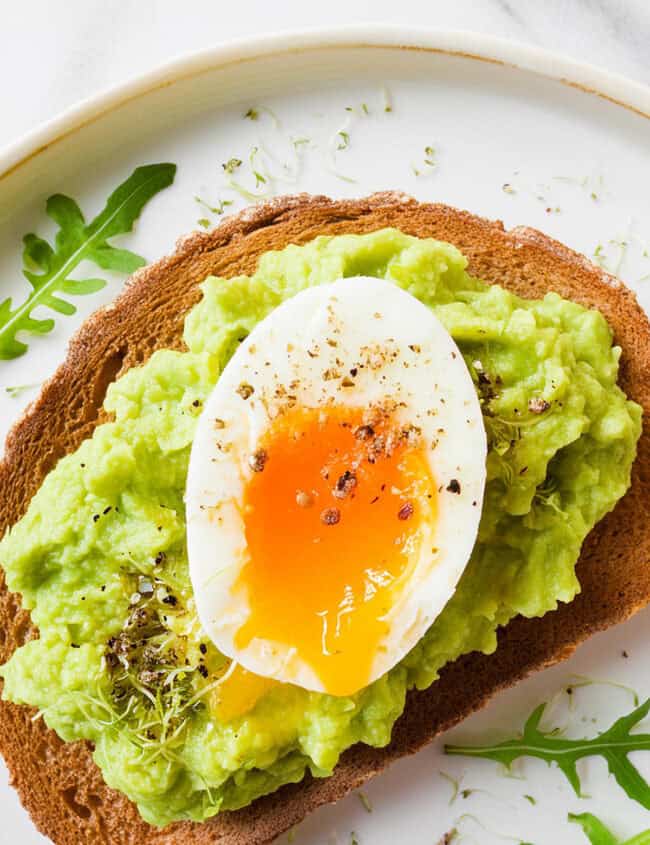 Enjoy Guacamole Toast: a slice of whole-grain toast topped with creamy mashed avocado and a runny, soft-boiled egg, garnished with freshly ground black pepper and sprinkled with microgreens. This delicious toast is served on a white plate with a few arugula leaves around the edges.