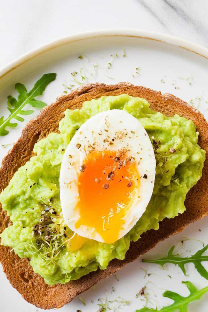 Enjoy Guacamole Toast: a slice of whole-grain toast topped with creamy mashed avocado and a runny, soft-boiled egg, garnished with freshly ground black pepper and sprinkled with microgreens. This delicious toast is served on a white plate with a few arugula leaves around the edges.