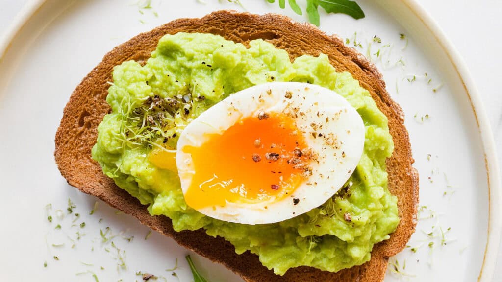 A plate with a slice of toasted bread topped with Guacamole and a halved soft-boiled egg, garnished with freshly ground black pepper and green herbs.