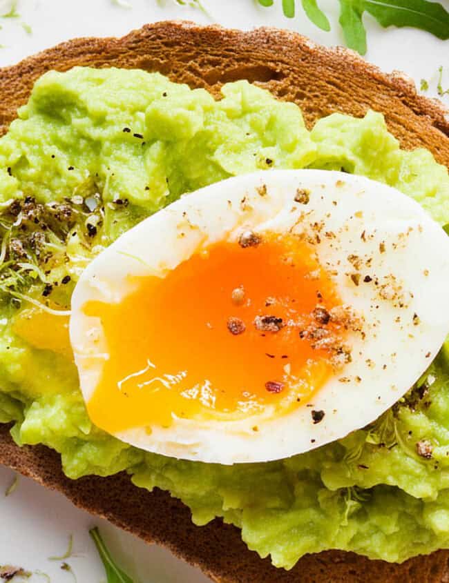 A plate with a slice of toasted bread topped with Guacamole and a halved soft-boiled egg, garnished with freshly ground black pepper and green herbs.