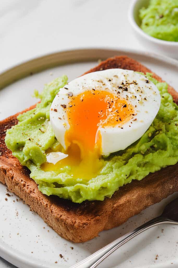 A slice of toasted bread topped with mashed avocado and a poached egg with a runny yolk, sprinkled with black pepper. The guacamole toast is on a white plate, with a bowl of Guacamole and a fork beside it.