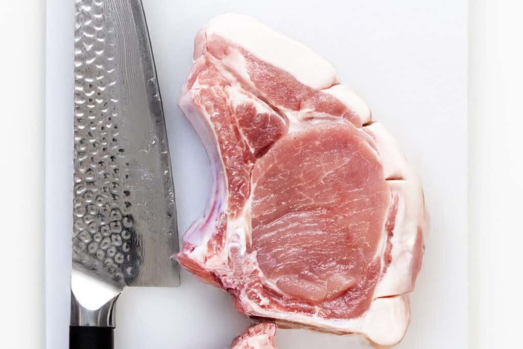 A raw pork chop, destined to become delectable smothered pork chops, rests on a white cutting board beside a large chef's knife with a textured metal blade and dark handle.