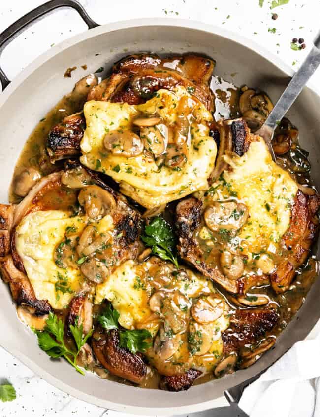 A pan of smothered pork chops topped with melted cheese, mushrooms, and a creamy sauce, garnished with parsley. A metal serving spoon rests in the pan.