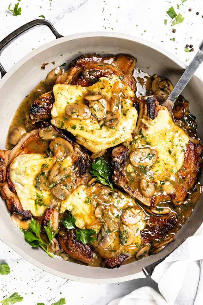 A pan of smothered pork chops topped with melted cheese, mushrooms, and a creamy sauce, garnished with parsley. A metal serving spoon rests in the pan.