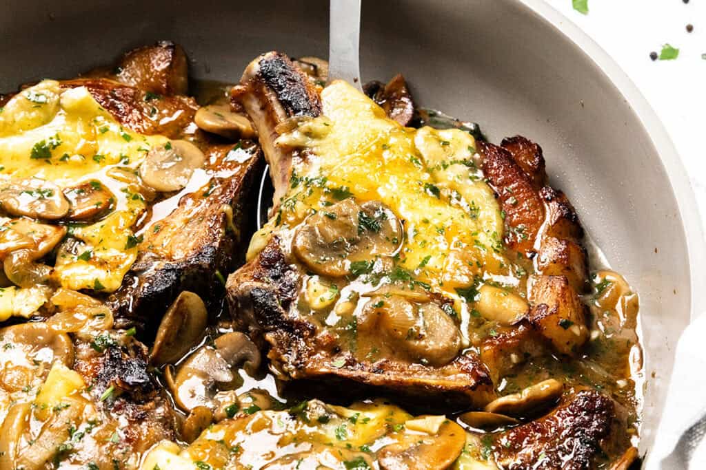 Close-up of smothered pork chops in a skillet, topped with melted cheese, sliced mushrooms, and herbs in a savory sauce. The dish appears golden and flavorful with visible seasoning.